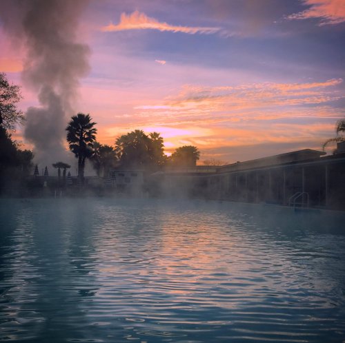 A serene pool scene at sunset, with steam rising and colorful clouds reflecting on the water's surface.