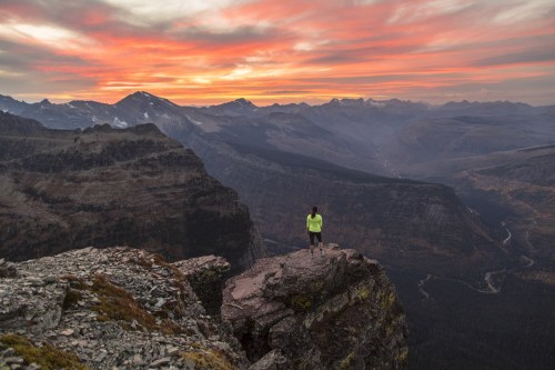 A person stands on a rocky ledge, overlooking a vast mountain landscape at sunset with vibrant skies.