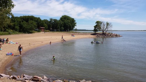 A sandy beach by a calm lake, with people swimming and relaxing under a clear blue sky. Trees line the shore.