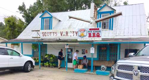 A small BBQ restaurant with a blue and white exterior, featuring a sign and people outside, surrounded by watermelons.