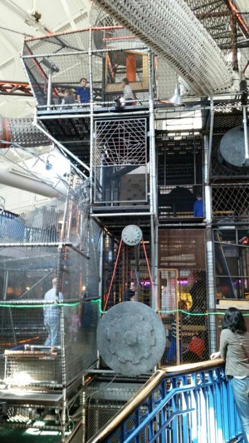 A large indoor play structure with multiple levels, nets, and climbing areas, filled with children playing and exploring.