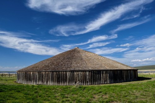 A round wooden structure with a conical roof, set against a blue sky with wispy clouds and green grass surrounding it.