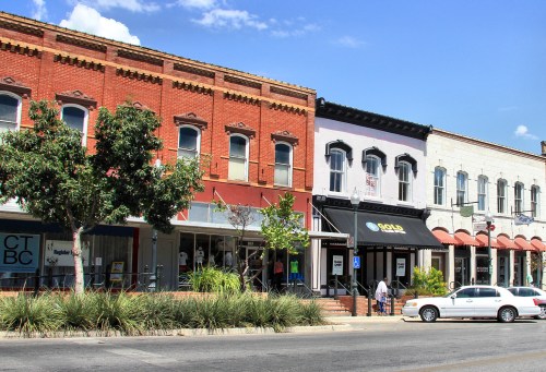 Historic storefronts line a sunny street, featuring red brick and white buildings with greenery and pedestrians.