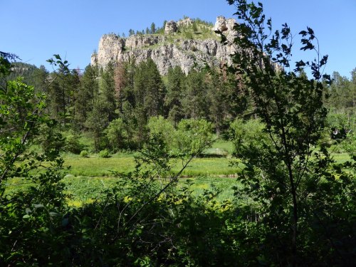 A rocky outcrop rises above a lush green landscape, surrounded by trees and a clear blue sky.