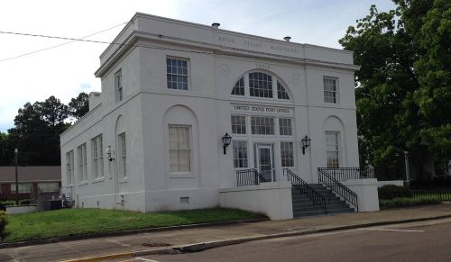 Historic white building with large windows, labeled "United States Post Office," surrounded by green grass and trees.