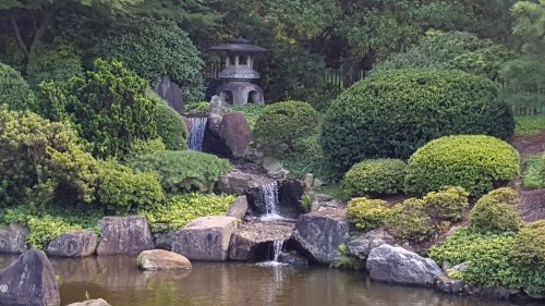 A serene garden scene featuring lush greenery, a waterfall, and a traditional pagoda beside a tranquil pond.