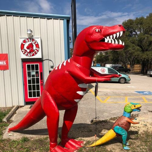 A large red dinosaur statue stands outside a building, with a smaller colorful dinosaur nearby on the ground.