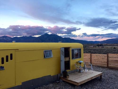 A bright yellow trailer with a wooden deck, set against a backdrop of mountains and a colorful sunset sky.