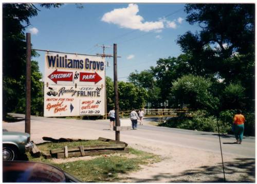 Sign for Williams Grove Speedway and Park, with people walking along a road under a blue sky and trees.