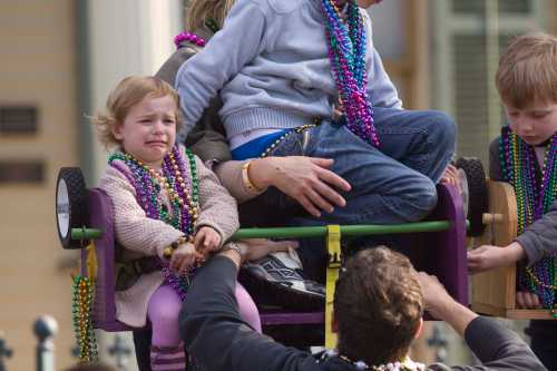 A child with a concerned expression sits in a wagon, surrounded by kids wearing colorful beads during a festive event.