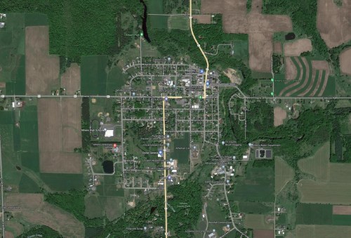 Aerial view of a small town surrounded by fields and forests, with roads and buildings visible.