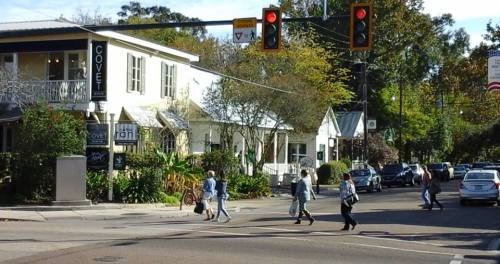 Pedestrians cross a street near shops and traffic lights in a suburban area with trees and parked cars.