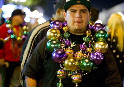 A man wearing a black shirt and a cap, adorned with colorful Mardi Gras beads, stands in a festive crowd at night.