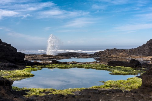 A rocky coastline with tidal pools reflecting the sky, and a water spout erupting in the background.