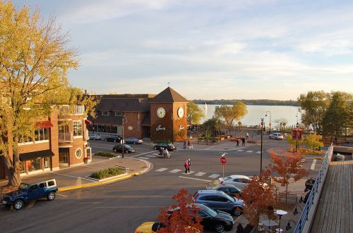A scenic view of a lakeside town square with shops, a clock tower, and autumn foliage.