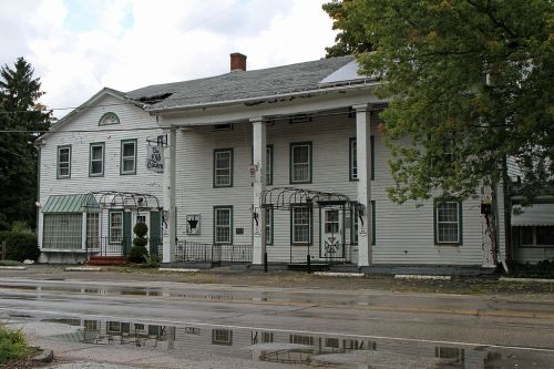 Historic white building with columns, large windows, and a porch, reflecting in a puddle on a rainy day.
