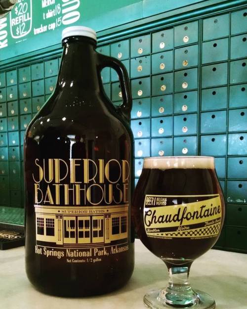 A large beer growler labeled "Superior Bathhouse" next to a glass of dark beer on a table.