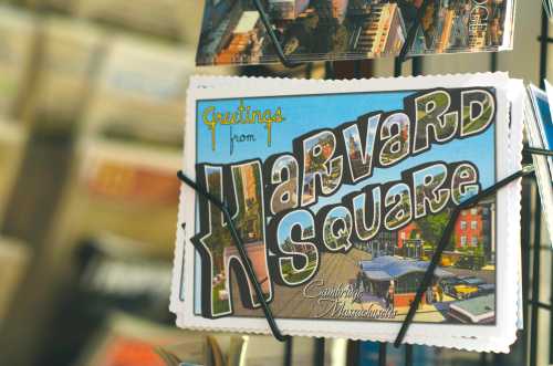 A colorful postcard reading "Greetings from Harvard Square, Cambridge, Massachusetts" displayed on a rack.