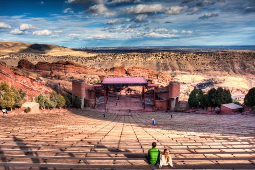 A couple sits on the steps of an outdoor amphitheater, overlooking a scenic landscape with mountains and clouds.