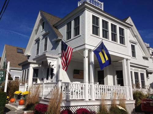 A white house with a porch, featuring American and rainbow flags, surrounded by colorful flowers and pumpkins.