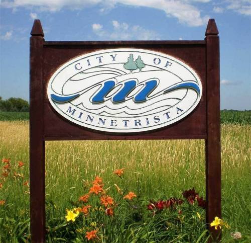 Sign for the City of Minnetrista, featuring a stylized "M" and surrounded by flowers and a grassy field.