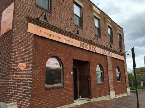 Brick building with a sign reading "Micucci Grocery" and "Formaggio-Vino" above the entrance. Cloudy sky in the background.