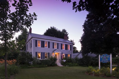 A charming two-story house with a welcoming porch, surrounded by lush greenery at dusk, with warm lights glowing from the windows.