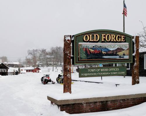 Sign for Old Forge Visitor Information Center in a snowy landscape, with snowmobiles parked nearby and an American flag.