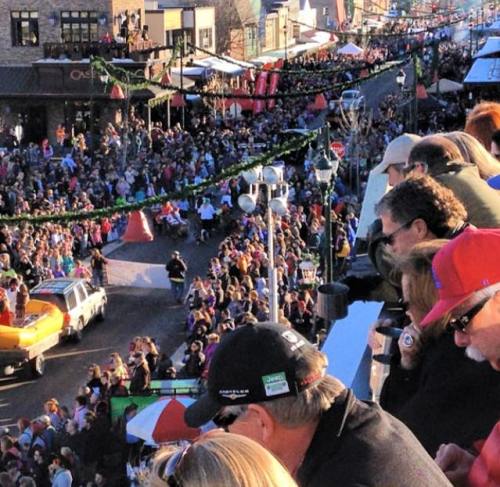 A large crowd watches a parade from a raised viewpoint, with festive decorations and colorful floats in the street below.