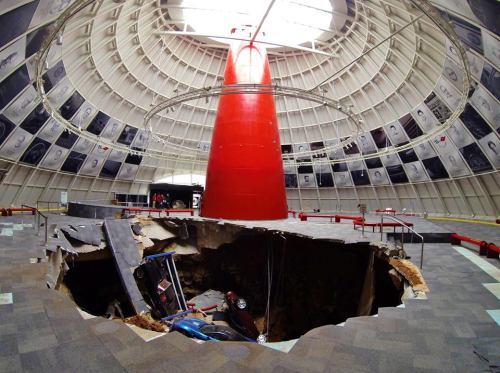 A large sinkhole in a modern, circular building with a red pillar, surrounded by posters and empty seating areas.
