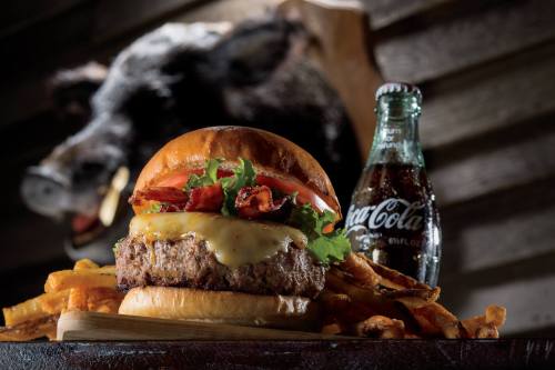 A juicy burger with cheese, lettuce, tomato, and bacon, served with fries and a bottle of Coca-Cola in the background.
