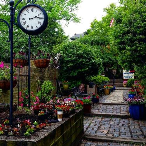 A charming garden path lined with colorful flowers, potted plants, and a clock on a post amidst lush greenery.