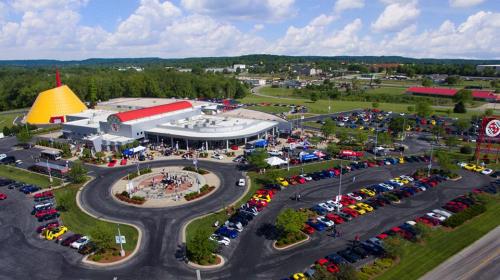 Aerial view of a busy restaurant with a colorful roof, surrounded by cars and green landscape under a blue sky.