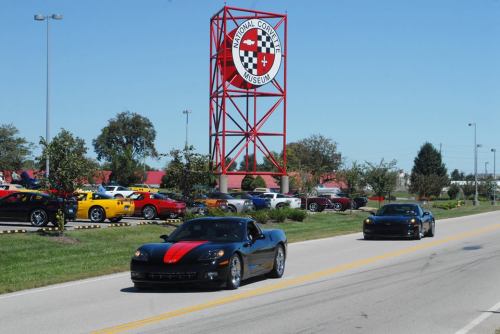 Two sports cars drive past the National Corvette Museum, with colorful Corvettes parked nearby under a clear blue sky.