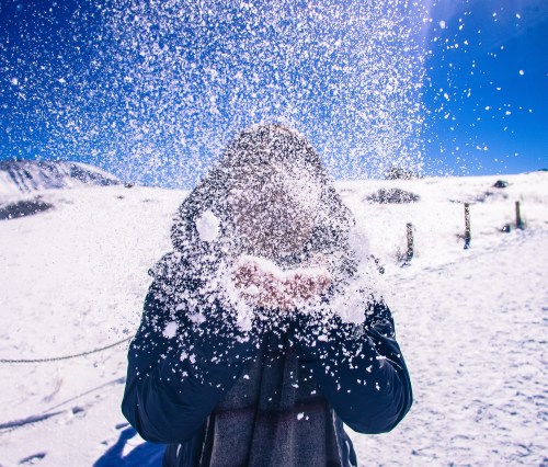 A person in a winter coat joyfully throws snow into the air against a bright blue sky and snowy landscape.