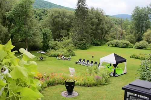 A lush green garden with lounge chairs, a tent, and flower beds, surrounded by trees and hills in the background.