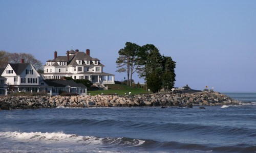A large, elegant house on a rocky shoreline with waves lapping at the beach under a clear blue sky.