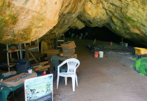 Interior of a cave with chairs, tools, and equipment, leading to a dark passageway.