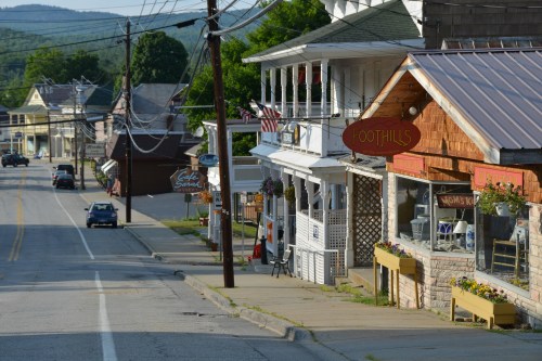 A quiet street in a small town featuring shops, a restaurant, and greenery in the background under clear skies.