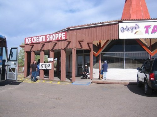 A rustic ice cream shop with a sign, featuring a few people outside and a bus parked nearby.