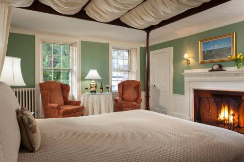 Cozy bedroom with a four-poster bed, two armchairs, a fireplace, and soft green walls. Natural light from windows.