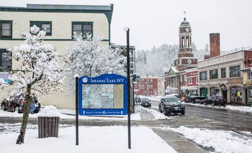Snow-covered street scene in Saranac Lake, NY, featuring a map sign and a clock tower in the background.