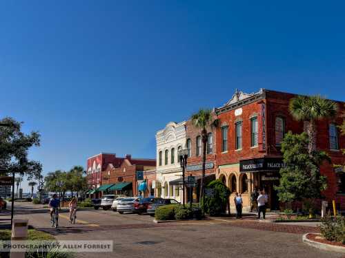 A sunny street scene featuring charming brick buildings, palm trees, and people walking and biking.