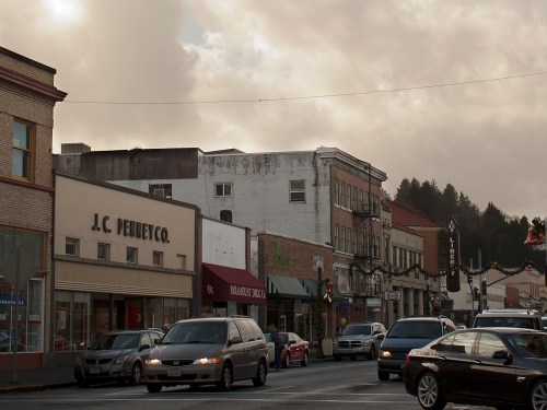 A street view of a small town with shops, cars, and festive decorations under a cloudy sky.