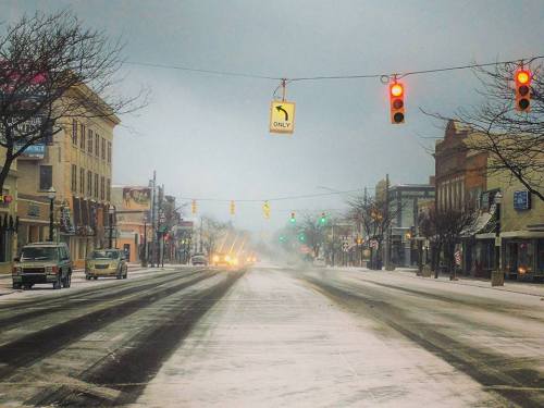 A snowy street scene with traffic lights, empty roads, and buildings lining the sides under a gray sky.