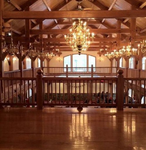 A spacious wooden hall with high ceilings, chandeliers, and a balcony overlooking a gathering below.
