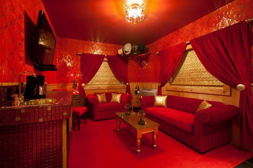 Cozy room with red walls, plush seating, gold accents, and a small bar area, illuminated by a chandelier.