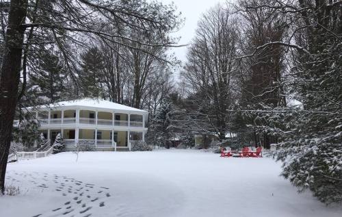 A snowy landscape featuring a large house and red chairs in a wintery yard surrounded by trees.