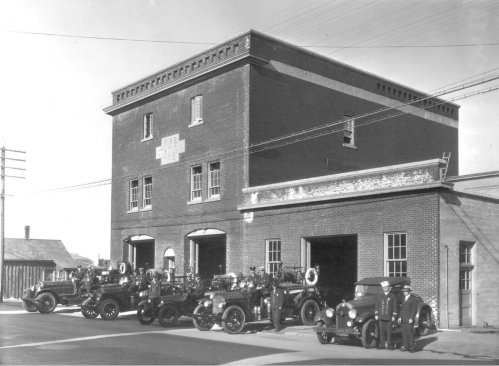 Historic black-and-white photo of a fire station with vintage fire trucks and firefighters in front.