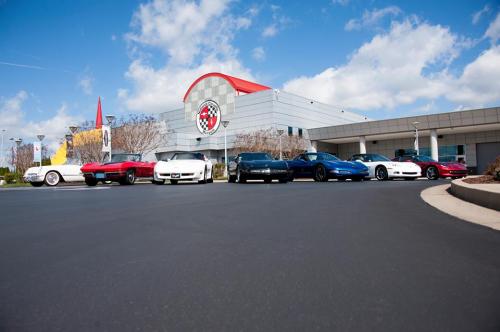 A lineup of classic Corvettes in front of a modern building with a red roof and a Corvette logo. Blue sky above.